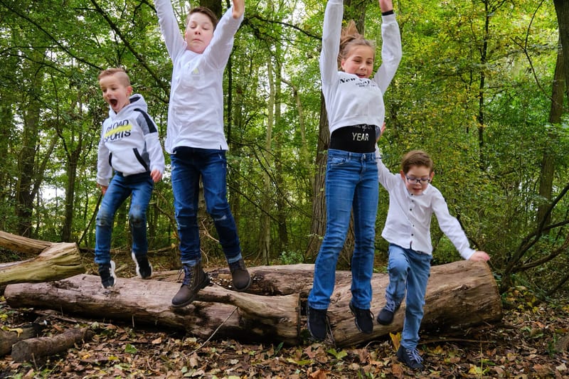 Vier kinderen springen blij op boomstammen in het bos, dynamische familiefotoshoot Westerpark Zoetermeer — Familieshoot Stefa